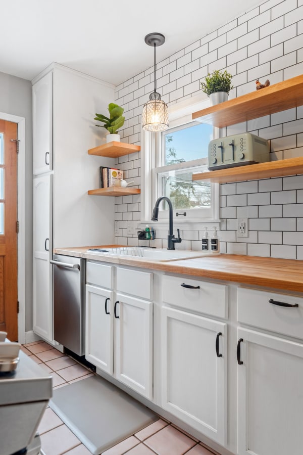 A bright kitchen with white cabinets, wooden countertops, and open shelves containing plants and cookbooks.