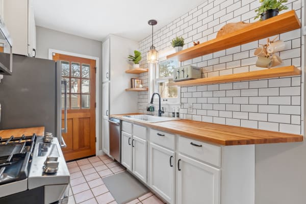 A cozy kitchen with a wooden countertop and white subway tile backsplash, featuring floating shelves and a glass pendant light.