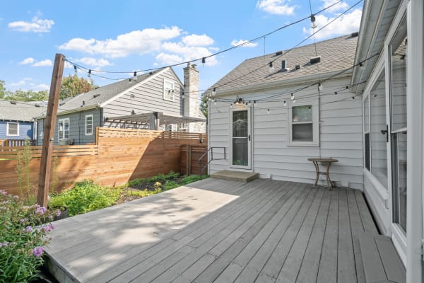 A deck with string lights and flowering plants, set against a blue sky and houses in the background.