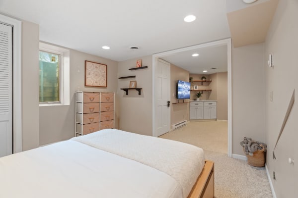 A cozy basement bedroom with a bed, storage unit, and a living area with a TV, all in neutral tones.