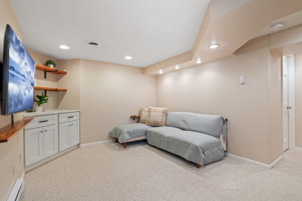 Cozy basement living area featuring a gray sectional couch, wall-mounted TV, and wooden shelves with plants.