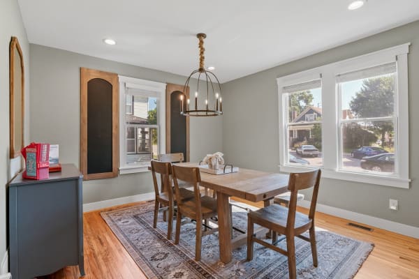 A cozy dining room featuring a wooden table, chairs, a chandelier, and large windows.