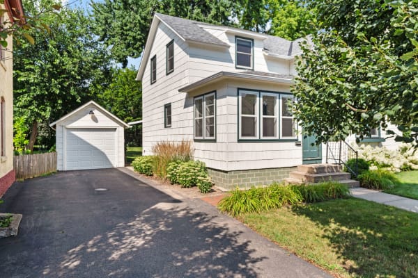 White two-story house with a dark green door and garage, surrounded by trees and gardens.