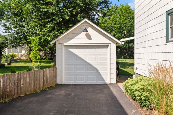 A white wooden garage with a closed door, surrounded by trees and garden plants.