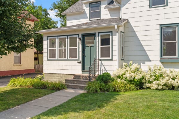 A two-story house with a white exterior and dark green accents, featuring a front porch and lush greenery.