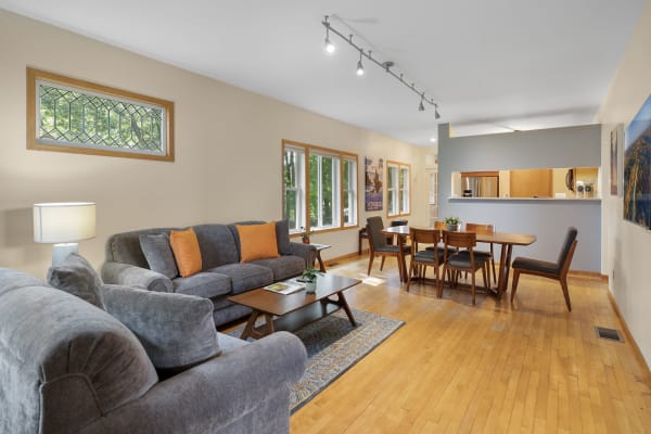 Living room with gray sofa, orange pillows, wooden dining table, and bright windows.