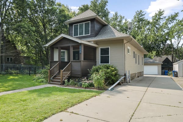 A two-story house with a porch, green lawn, and driveway, set in a residential area with trees.