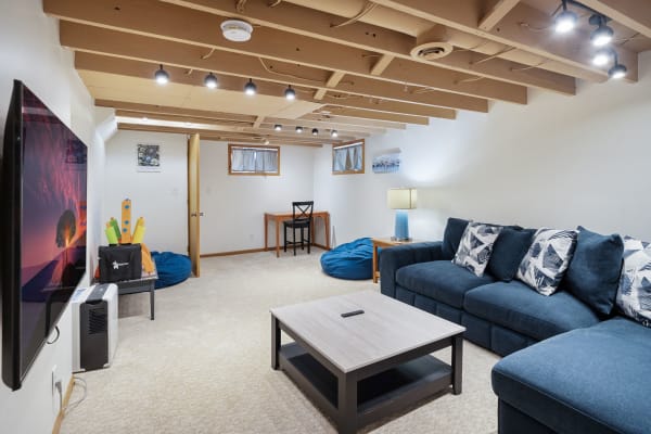 A well-lit basement living room featuring a blue sectional sofa, a small desk, and bright decor.