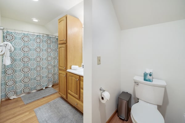 A neat bathroom featuring a patterned shower curtain, wooden cabinets, and a toilet area with tissues and a trash bin.