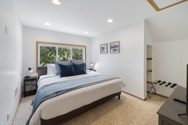 A modern bedroom featuring a queen-sized bed with navy-blue pillows, a black bedside table, and natural light from a large window.