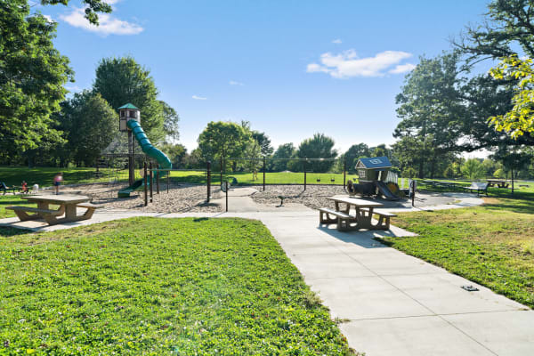 A playground with a slide, sandbox, swings, and a playhouse surrounded by green trees.