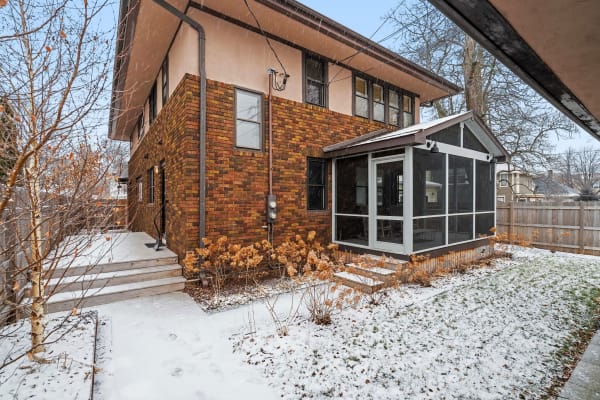 Exterior side view of a two-story house with brick and plaster, a snowy yard, and a screened porch.