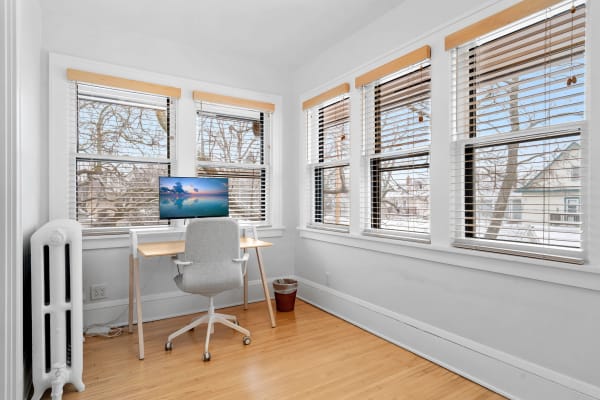 A well-lit office space featuring a wooden desk with a monitor, a gray chair, and large windows with white blinds.