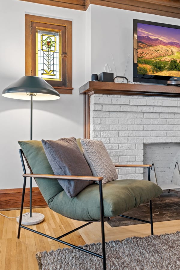 A cozy living room with a green lounge chair, stained glass window, and a TV above a white fireplace.