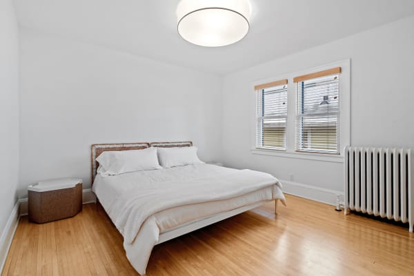 A serene and minimalist bedroom with a double bed, wooden floor, and natural light from the windows.