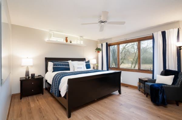 A cozy bedroom featuring a dark bed frame, white bedding, and a view of trees through a large window.