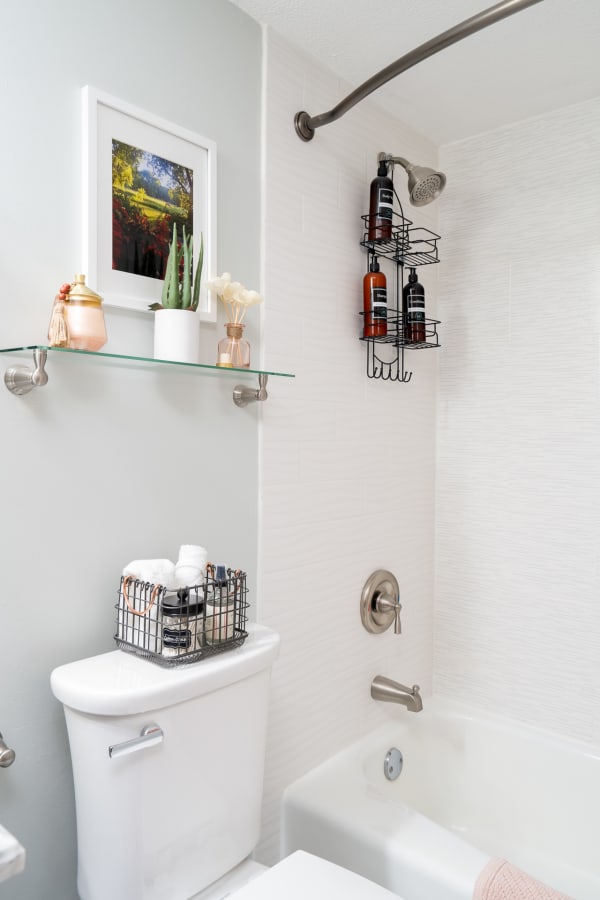A stylish bathroom with a gray wall, white tiles, a glass shelf, and decorative items featuring a bathtub and wire rack for toiletries.