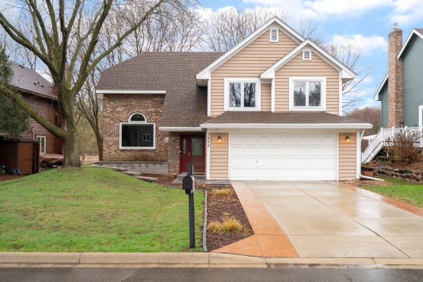 A two-story house with a brick and beige siding exterior, featuring a lush green yard and concrete driveway.