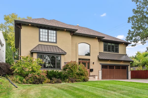 Modern two-story house with beige stucco and a dark roof, surrounded by green grass and mature plants.