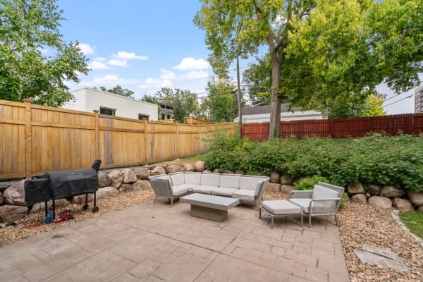 A contemporary backyard with a sectional sofa, coffee table, and grill surrounded by greenery and a wooden fence.
