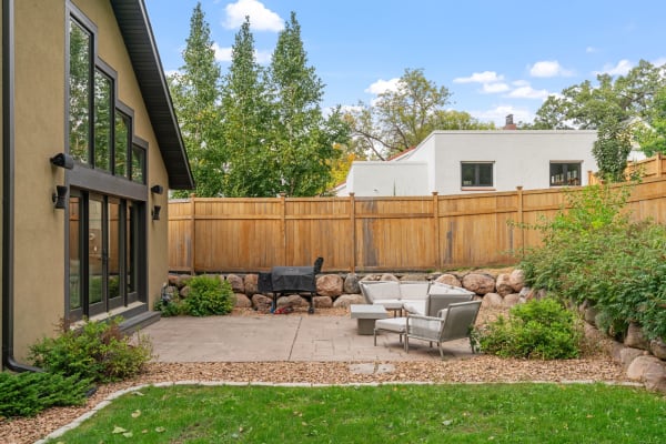 A modern patio with seating and a grill, surrounded by a wooden fence and trees.