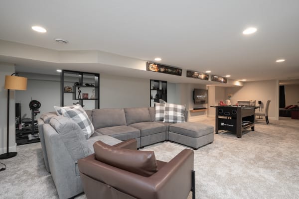 A cozy basement with a gray sectional sofa, foosball table, and decorative shelves.
