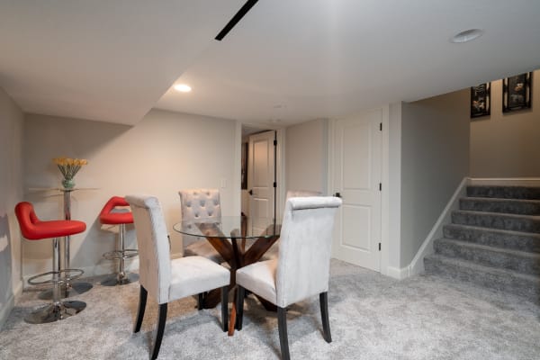A modern basement space with a round glass dining table and plush chairs, accentuated by red bar stools and a staircase in the background.