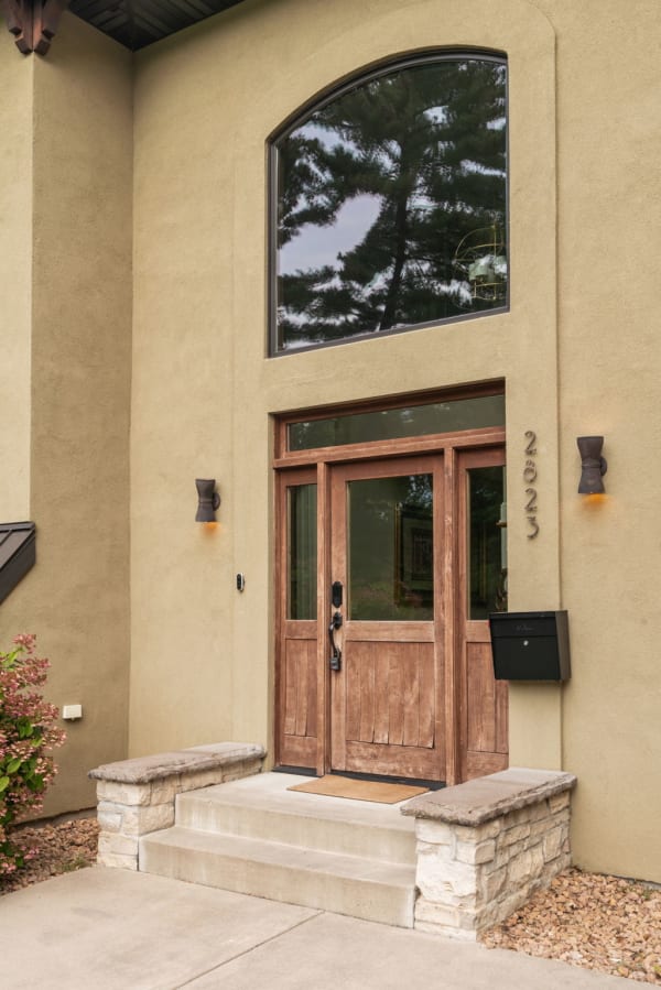 Modern home entrance with wooden doors and arched window surrounded by greenery.