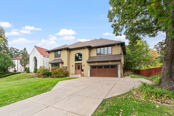 A modern two-story house with a tan exterior and a spacious driveway, surrounded by greenery and a clear sky.