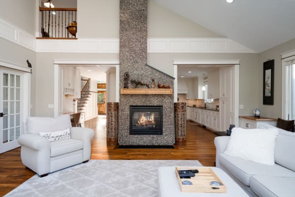 A modern living room featuring a stone fireplace, white furniture, and an open kitchen view.