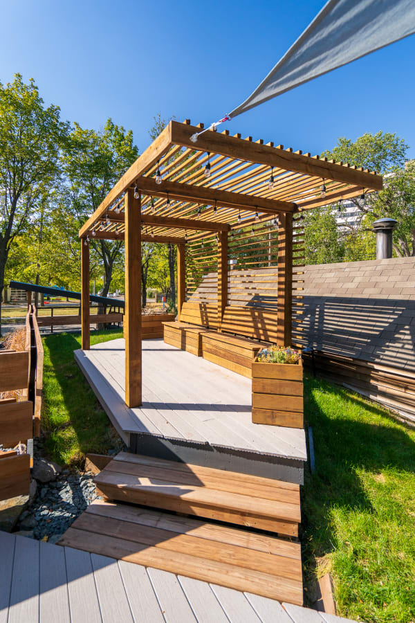 An outdoor pergola with a wooden deck and planters, set in a green landscape.
