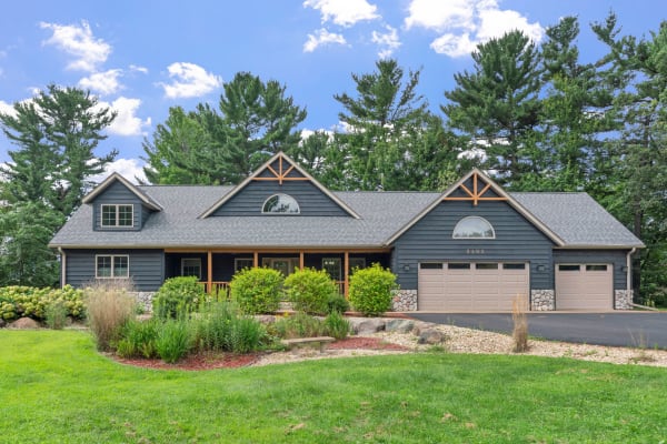 A two-story blue house with a front porch and garden, set amidst trees and blue sky.