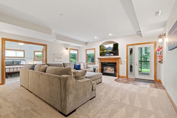 A cozy living room with a sectional sofa, fireplace, and large windows overlooking greenery.
