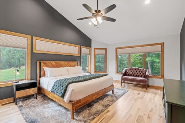 Modern bedroom with a bed, nightstand, and tufted loveseat, featuring large windows and wooden flooring.