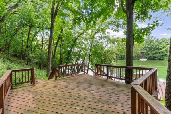 Wooden deck by a lake with lush trees and a sailboat moored in the background.
