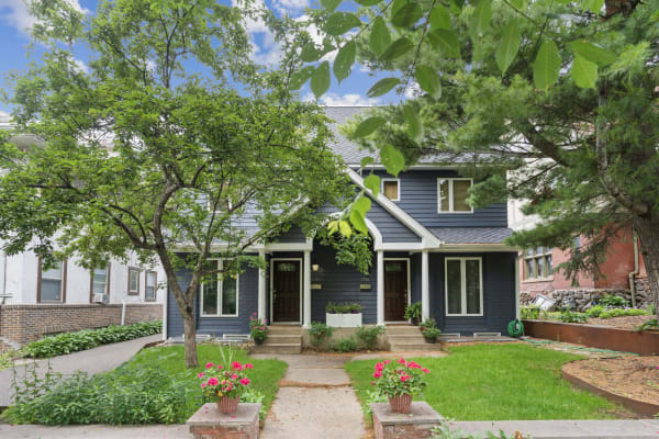 A two-story dark blue house with white trim, surrounded by greenery and pink flowers in pots.