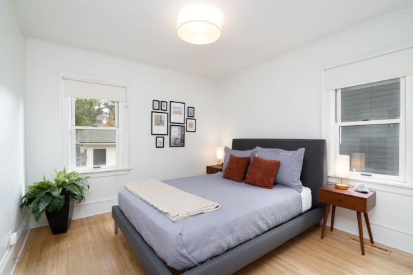 A bright bedroom with a gray bed, rust-colored pillows, a wooden nightstand, and framed artworks.