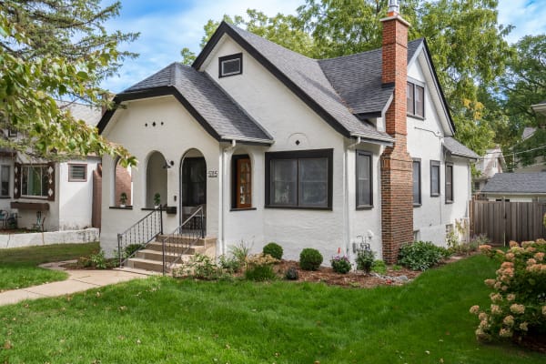 Exterior view of a charming white house with dark trim, featuring arched doorways, a brick chimney, and green landscaping.