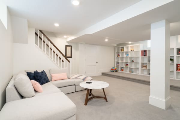 Bright basement living area featuring a light gray sectional sofa and white shelving with toys and books.