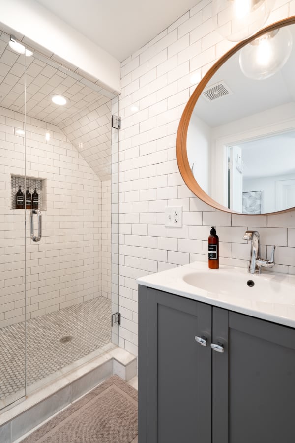 Modern bathroom featuring white subway tiled walls, a glass shower enclosure, and a round wooden-framed mirror above a sink.