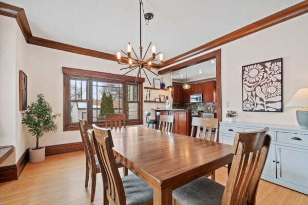 A dining room featuring a wooden table, chairs, and a modern chandelier with an adjoining kitchen visible.