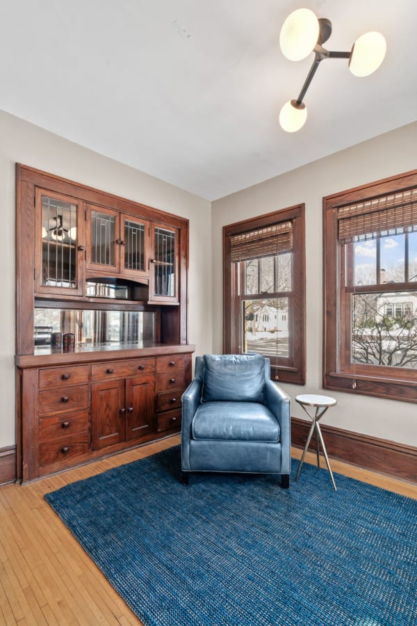 A corner of a room with a blue leather chair, wooden cabinet, and large windows.
