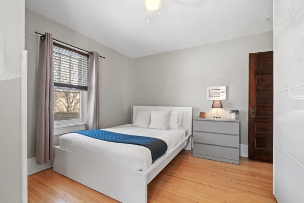 A bright and tidy bedroom featuring a bed with white linens and a blue quilt, a gray dresser, and a window with gray curtains.