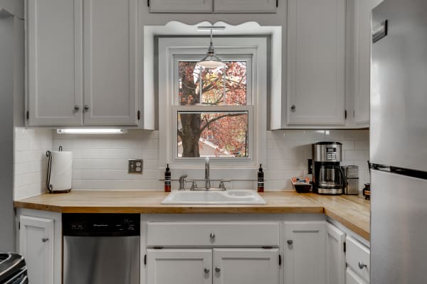 A modern kitchen with white cabinets, wooden countertop, and a view of autumn leaves through the window.