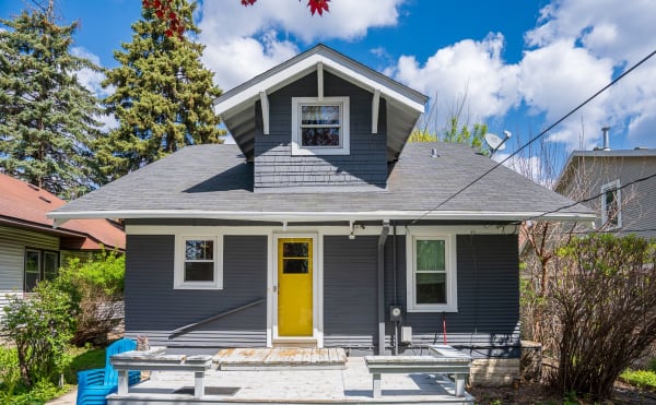 A gray house with a yellow door and blue chairs on the front deck, surrounded by trees.