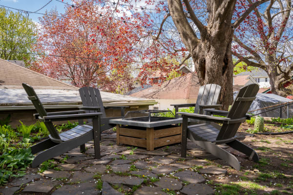 A cozy backyard scene featuring Adirondack chairs surrounding a fire pit, with trees and rooftops in the background.