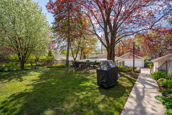 A green backyard with flowering trees, outdoor seating, and a covered grill.