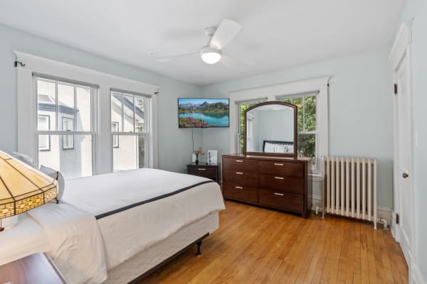 Spacious bedroom with large windows, a bed, a dresser, and a TV mounted on the wall.