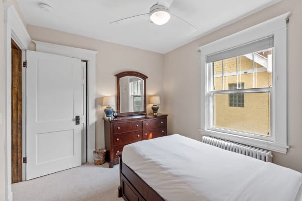 A cozy bedroom with a white bed, wooden dresser and mirror, and a window showing a view of an exterior wall.