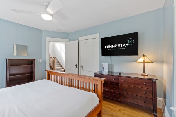 A cozy bedroom featuring a wooden bed frame, dresser, and modern ceiling fan, with a flat-screen TV on the wall.